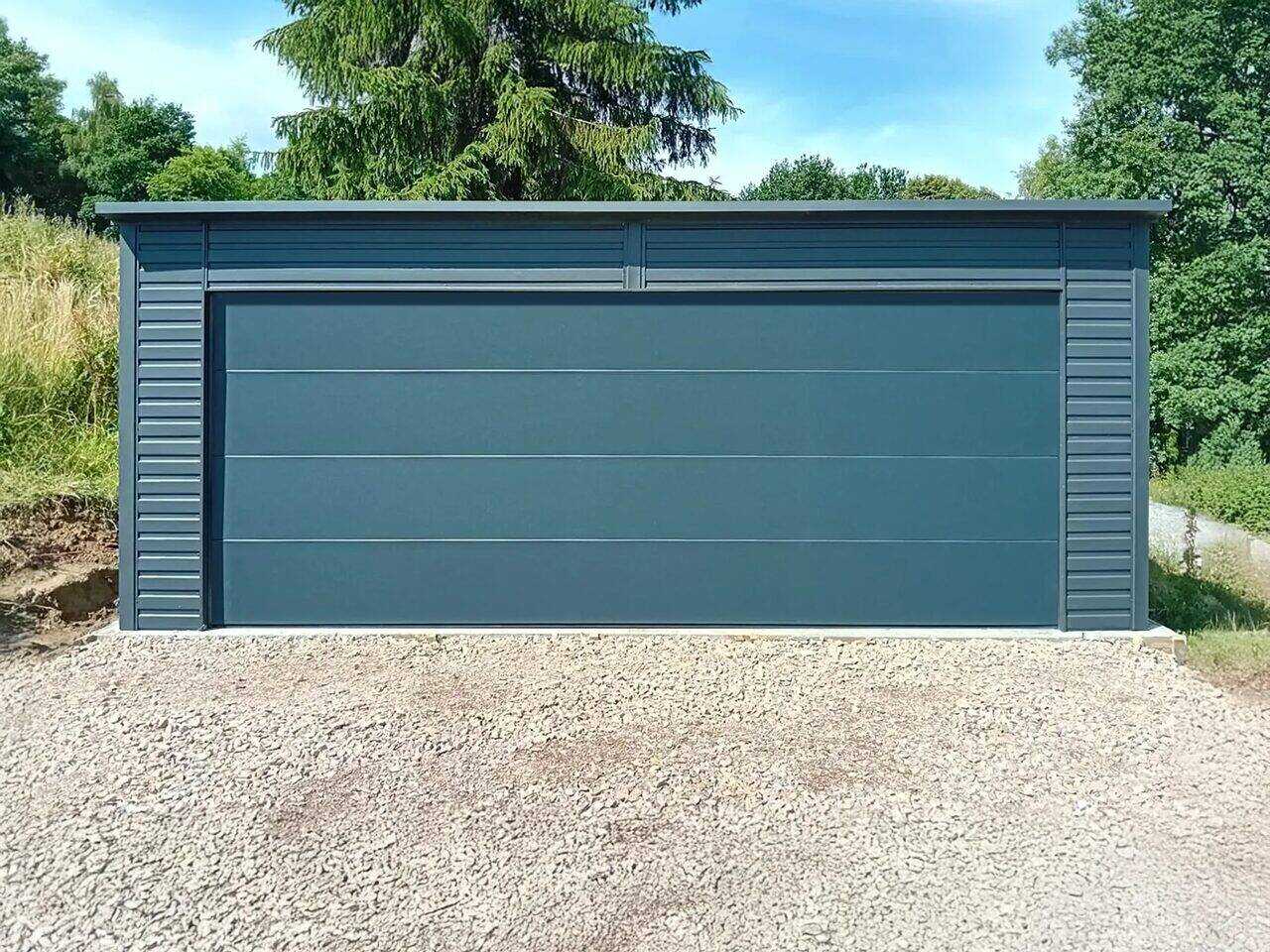 Anthracite steel garage with closed sectional door, straight front view on gravel with trees behind