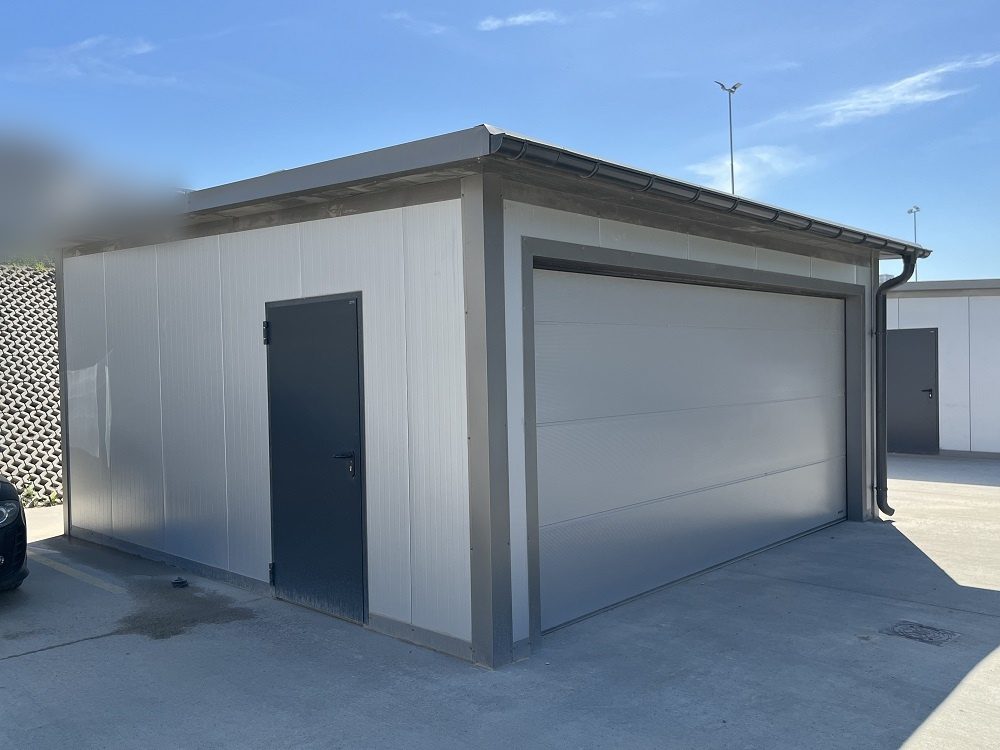 Insulated garage in grey panels with one wide light-grey sectional door and a side entrance door, on a paved yard