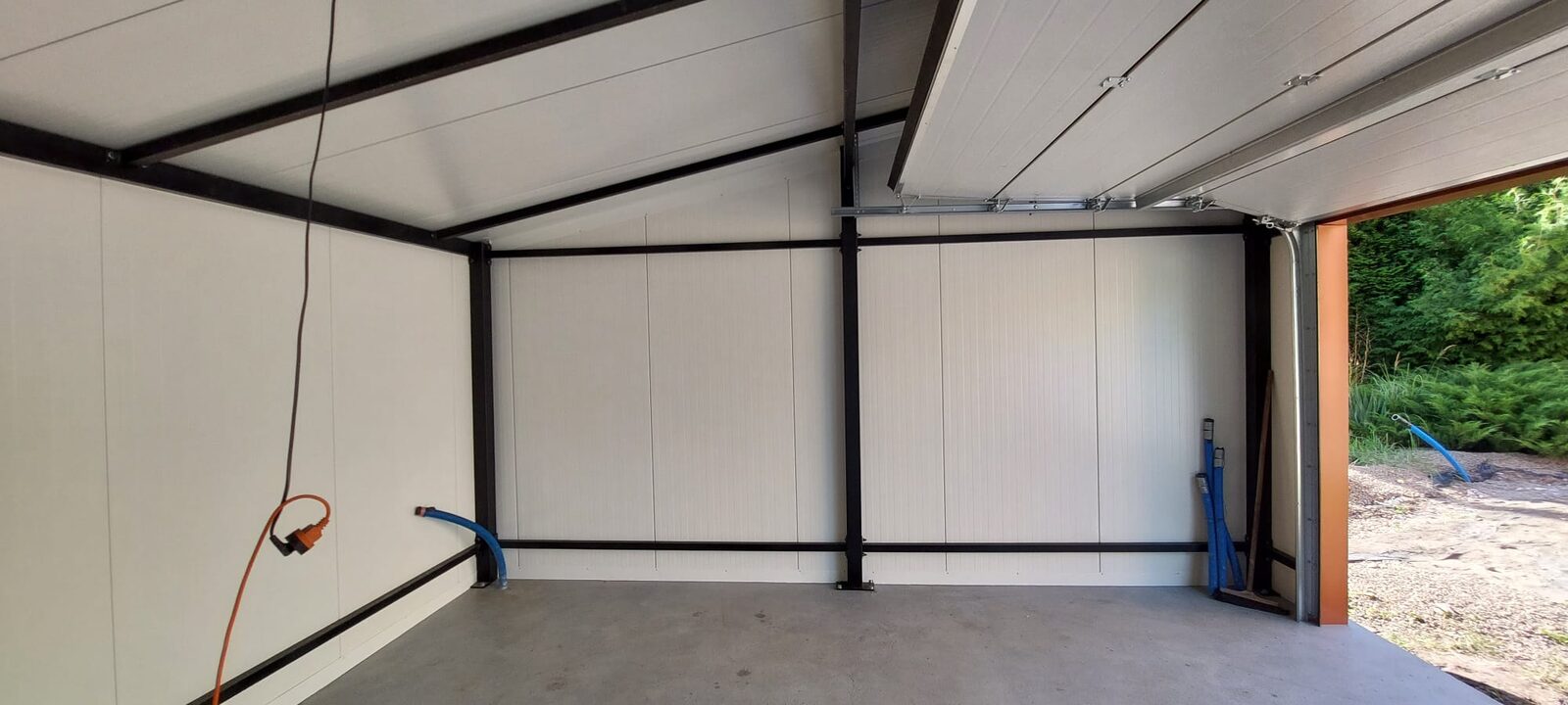 Wide interior view of an insulated garage roof apex, orange power cable and blue hose along the wall, open door onto greenery