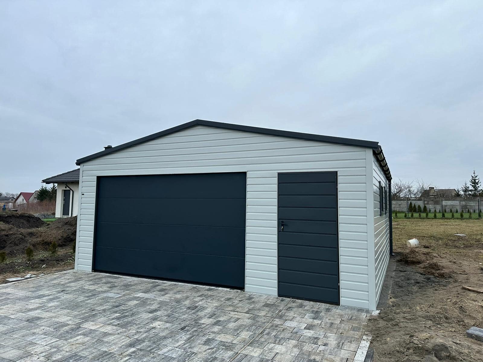 Light-grey steel garage with one anthracite sectional door beside a side entrance door, on a block-paved drive