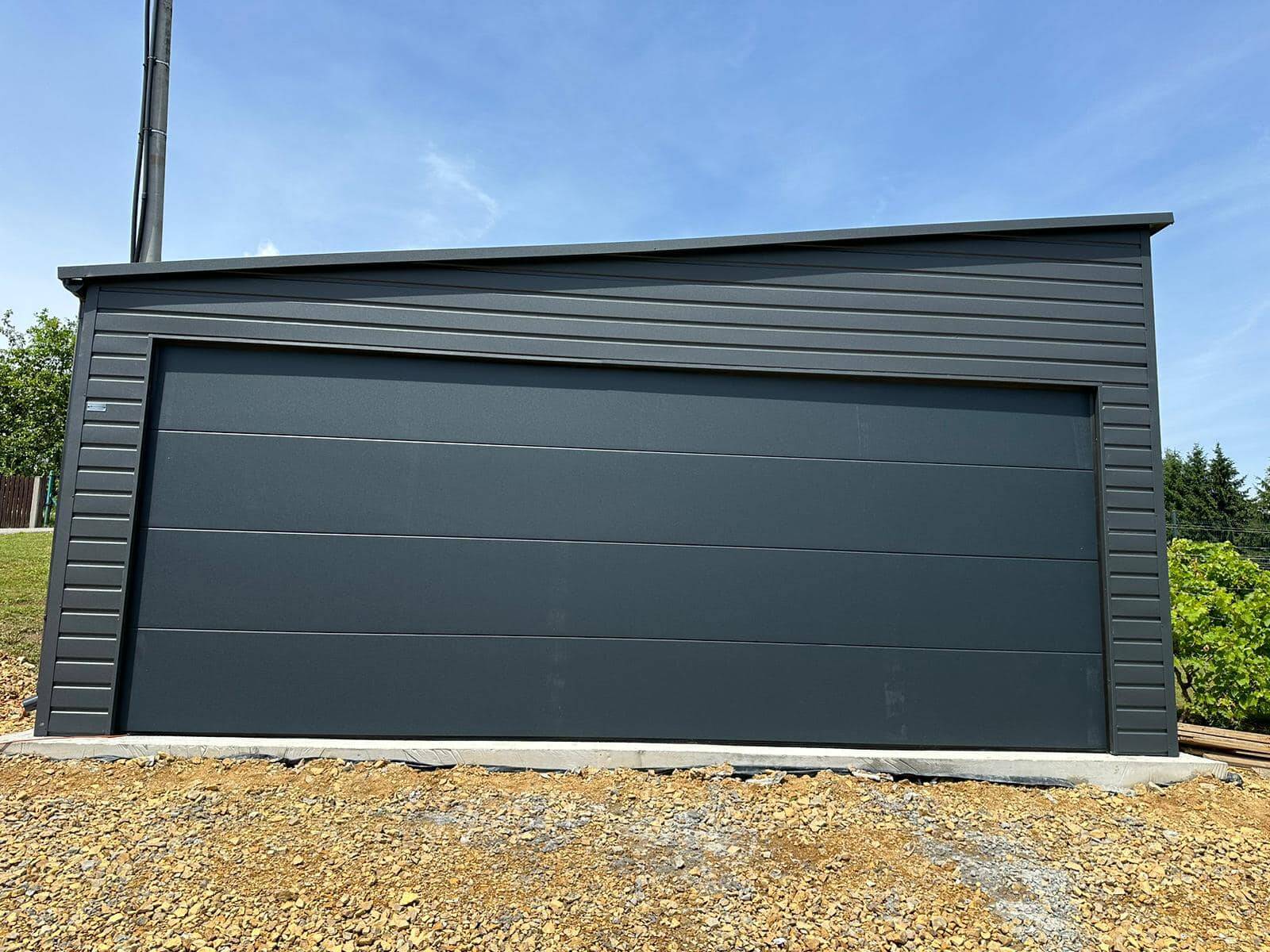 Anthracite steel garage with one wide sectional door and a roof sloping to one side, on a freshly laid base