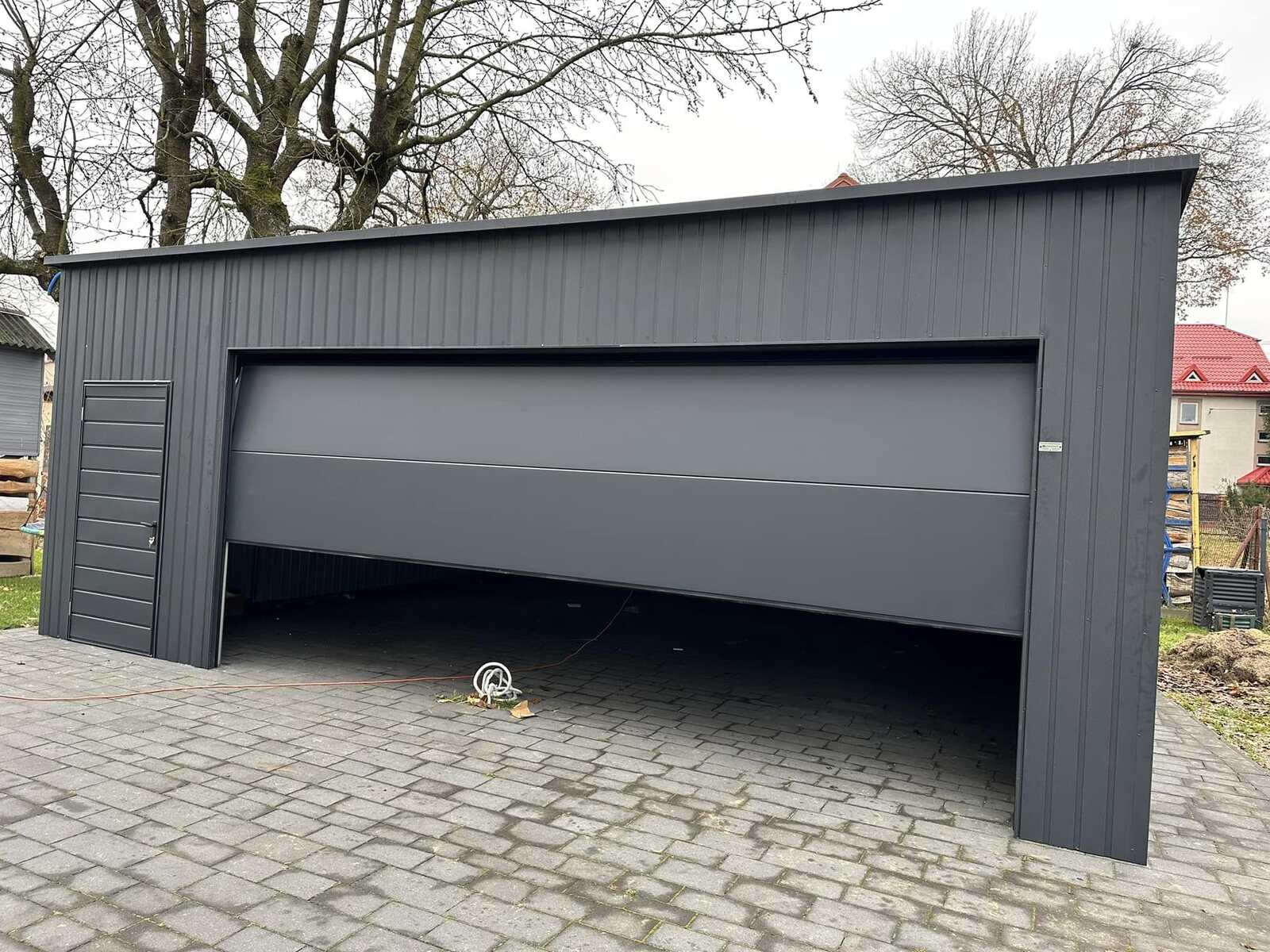 Anthracite steel garage with its wide door half raised and a side entrance door, on block paving