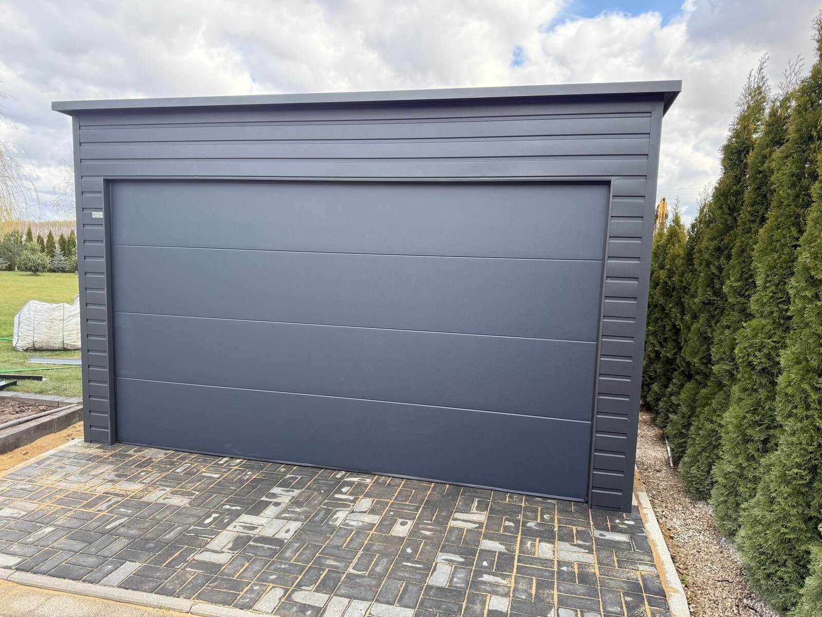 Anthracite steel garage with one wide sectional door, framed by conifers, on a block-paved apron