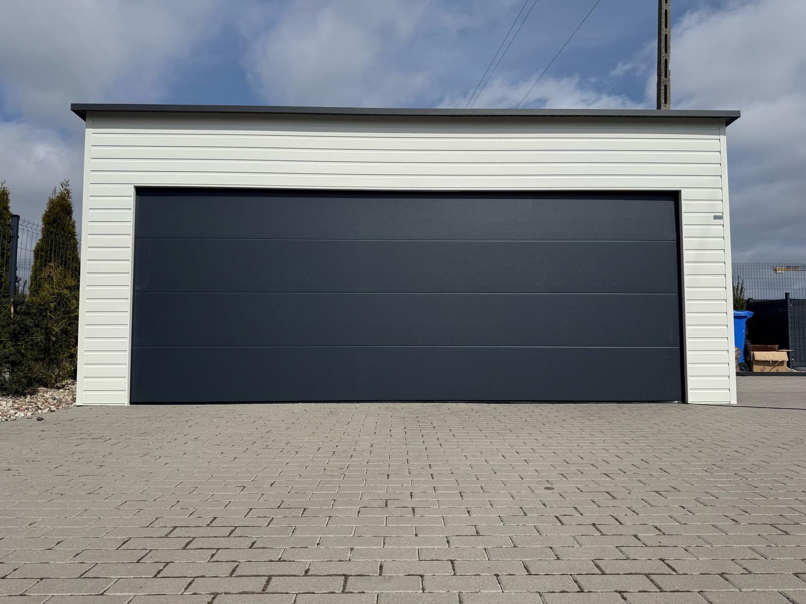 White-clad steel garage with a wide anthracite sectional door and a low roof, on a block-paved drive