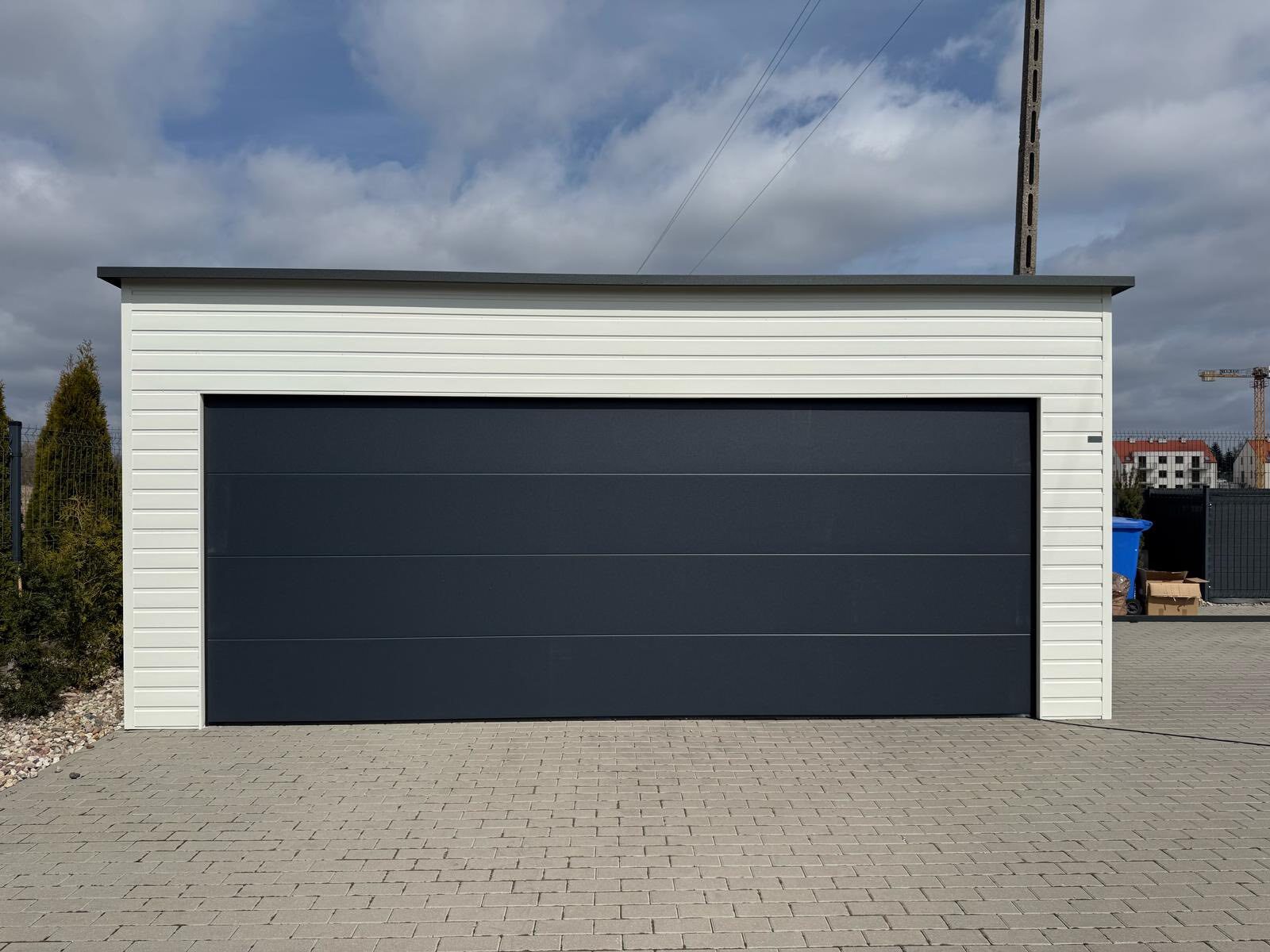 White-clad steel garage seen straight on across a paved yard, one wide anthracite sectional door