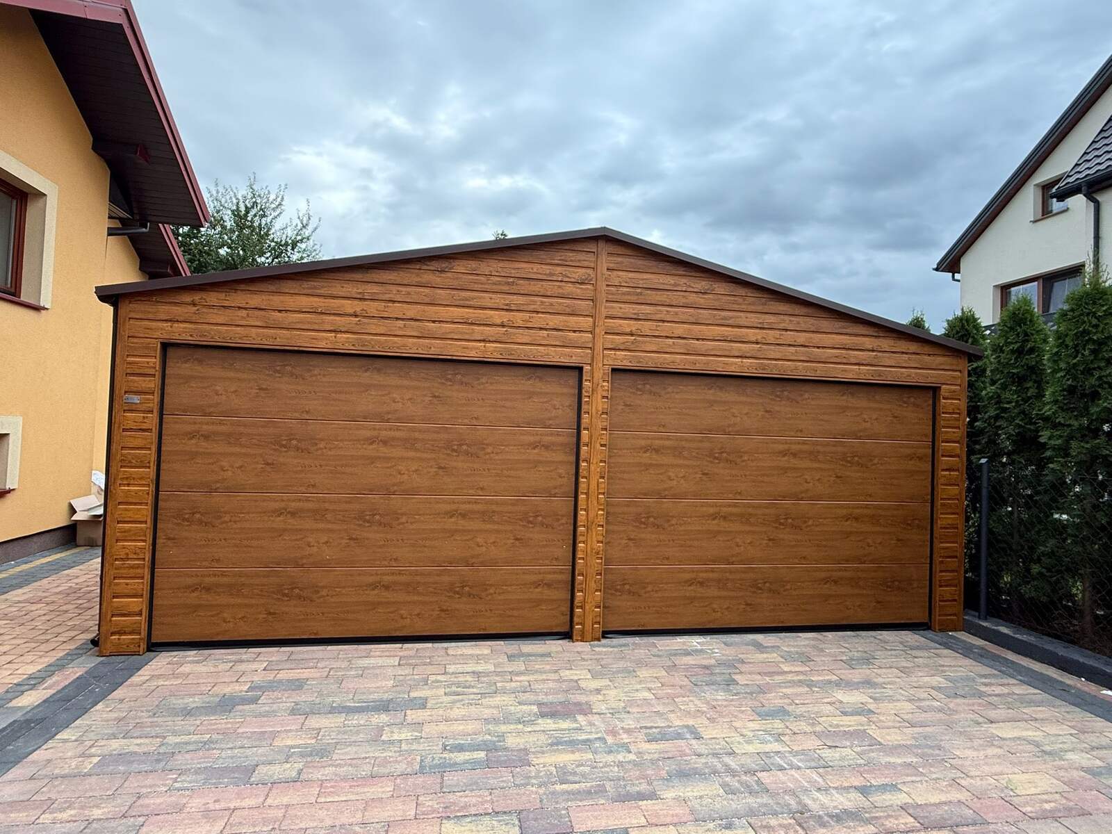 Wood-effect steel garage seen from the front, two sectional doors under an apex roof, between two houses