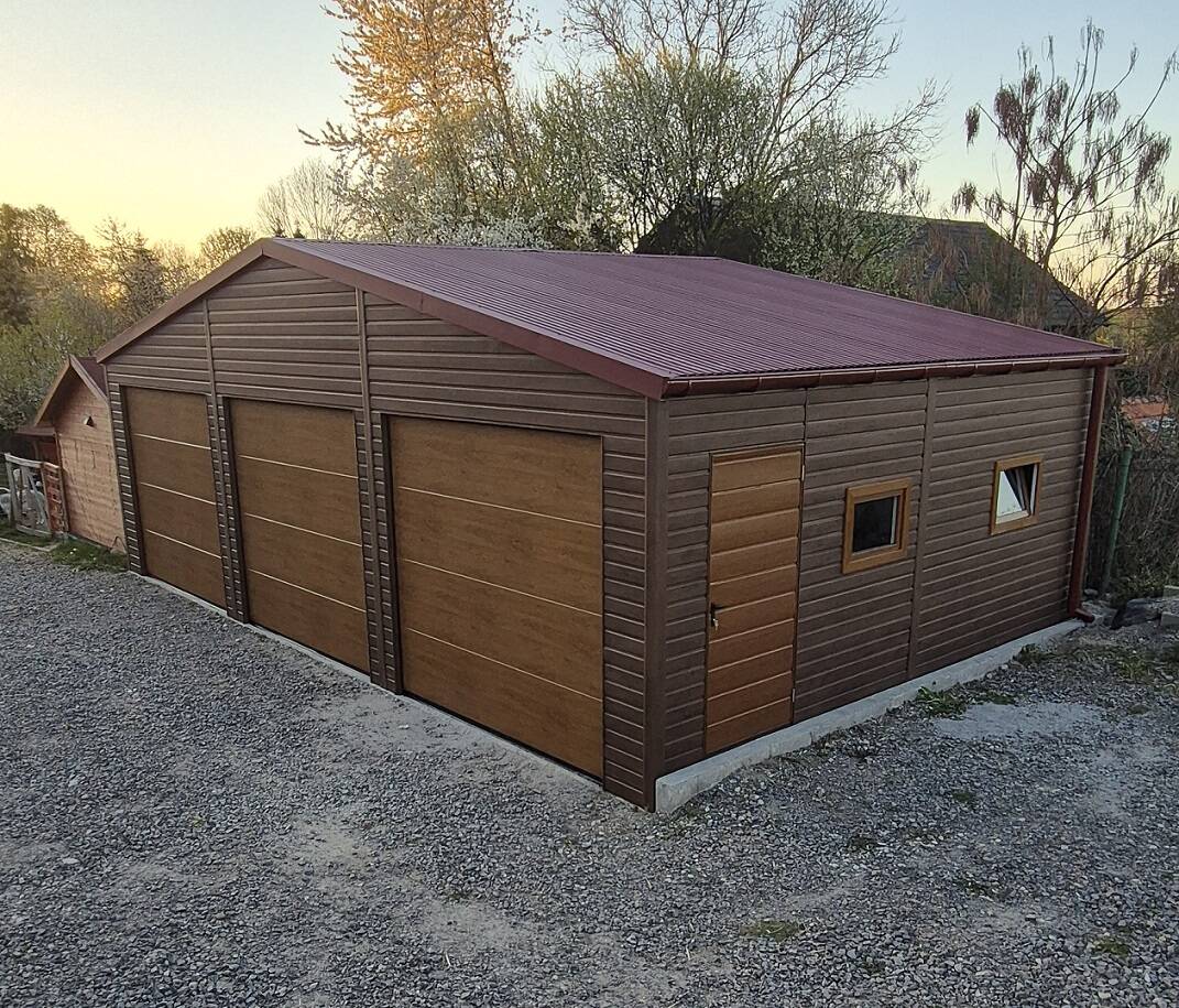 Brown steel garage with a red apex roof, three wood-effect sectional doors, a side entrance door and two windows, at dusk